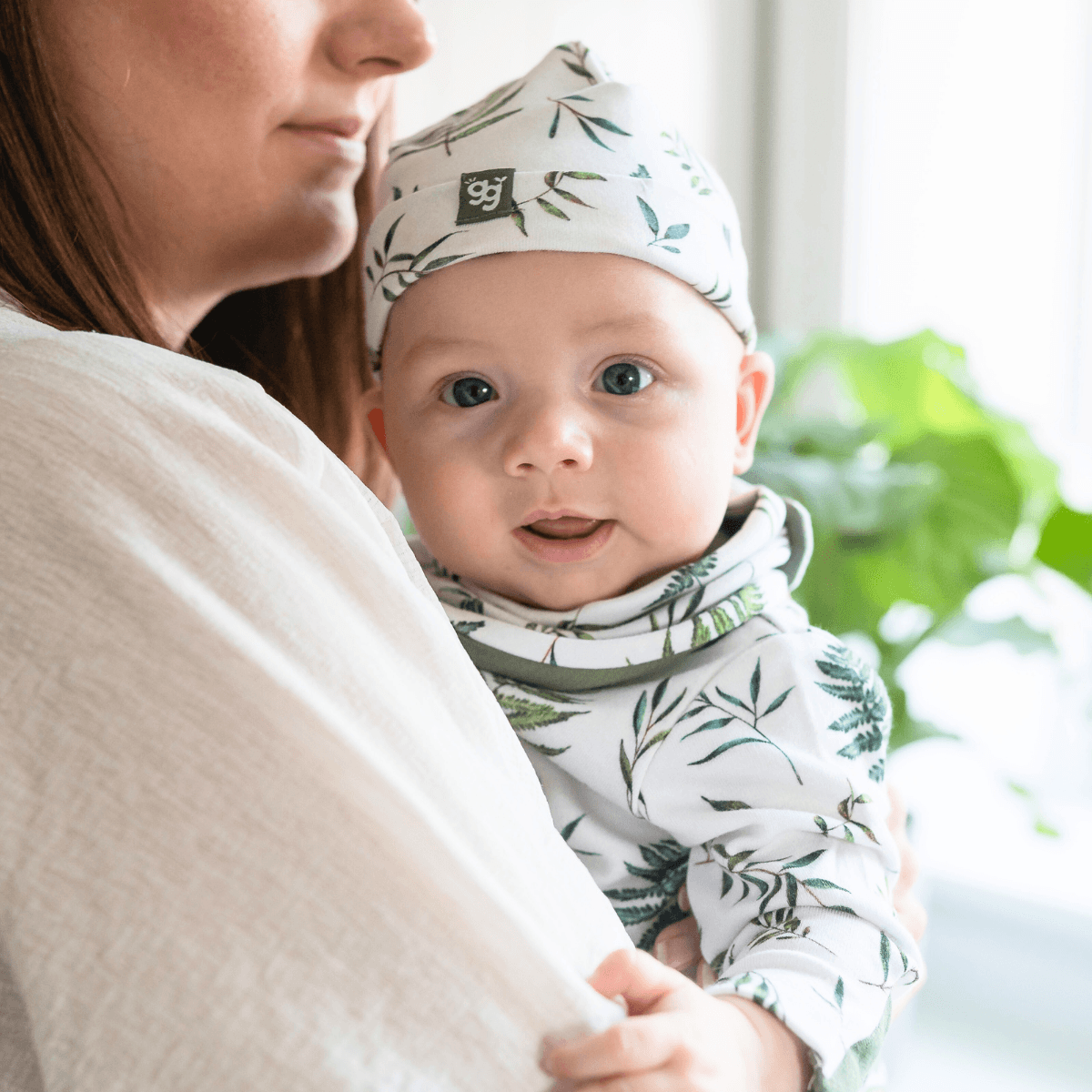 A baby wearing a matching GrowGrows sleepsuit with a matching baby hat and baby bib, held by an adult in a soft cream top. The background is a bright, softly blurred indoor setting with green foliage.