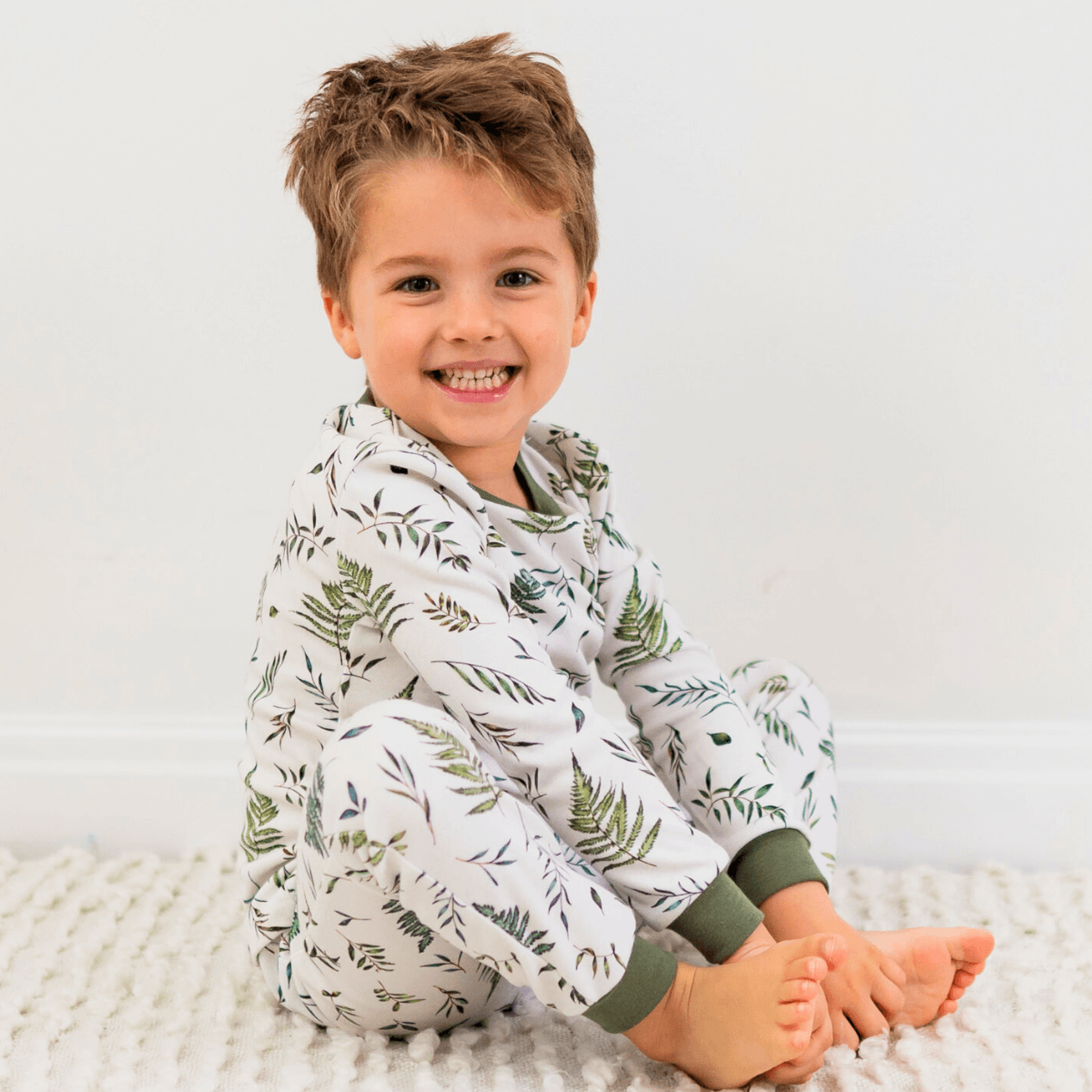 Child sat wearing the Country Leaf Pyjamas with green botanical leaf print on a white background made from bamboo and organic cotton from GrowGrows