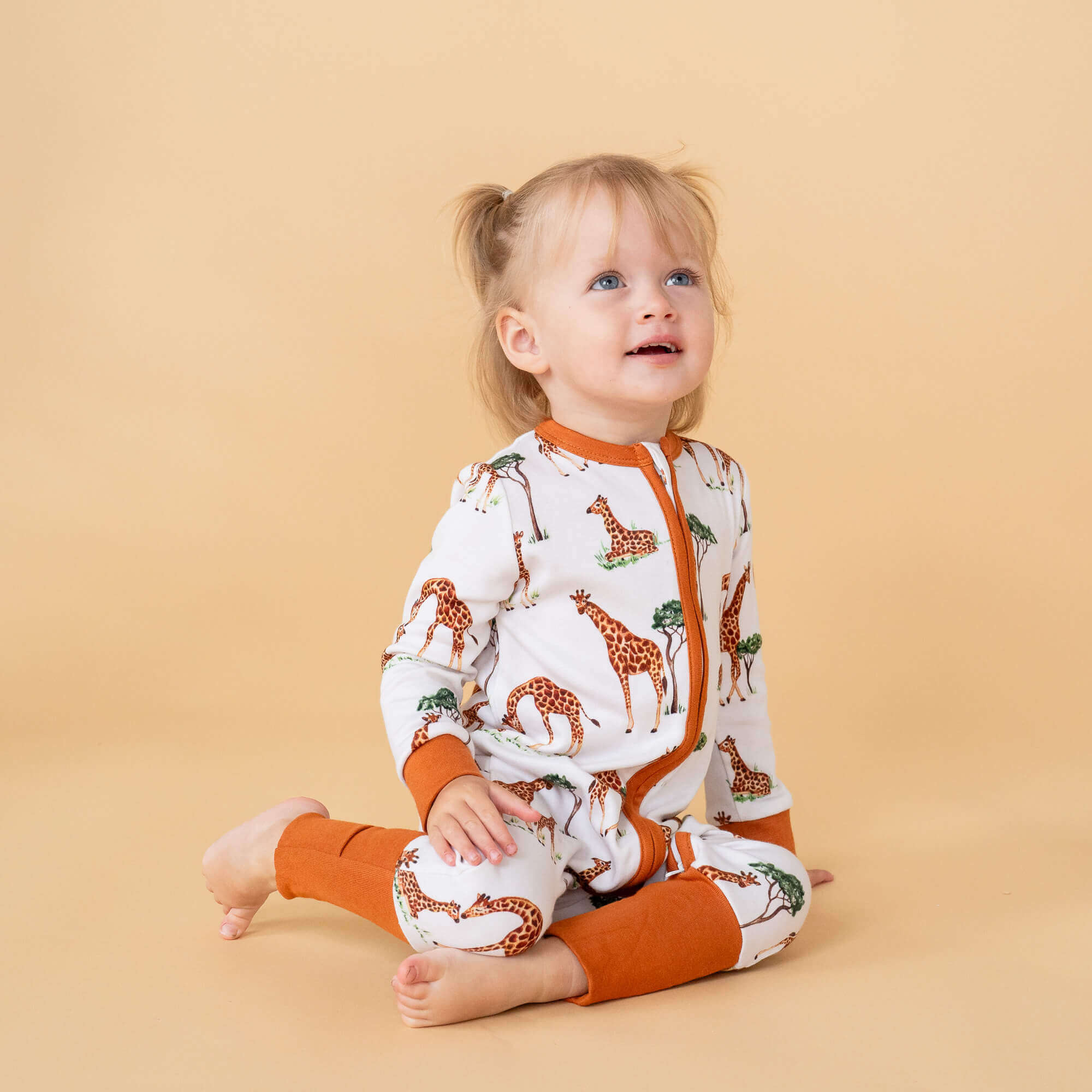 A toddler sitting on the floor with an orange background wearing the Gentle Giraffes Sleepsuit with mum and baby giraffe savannah print and desert orange trim made from bamboo and organic cotton from GrowGrows
