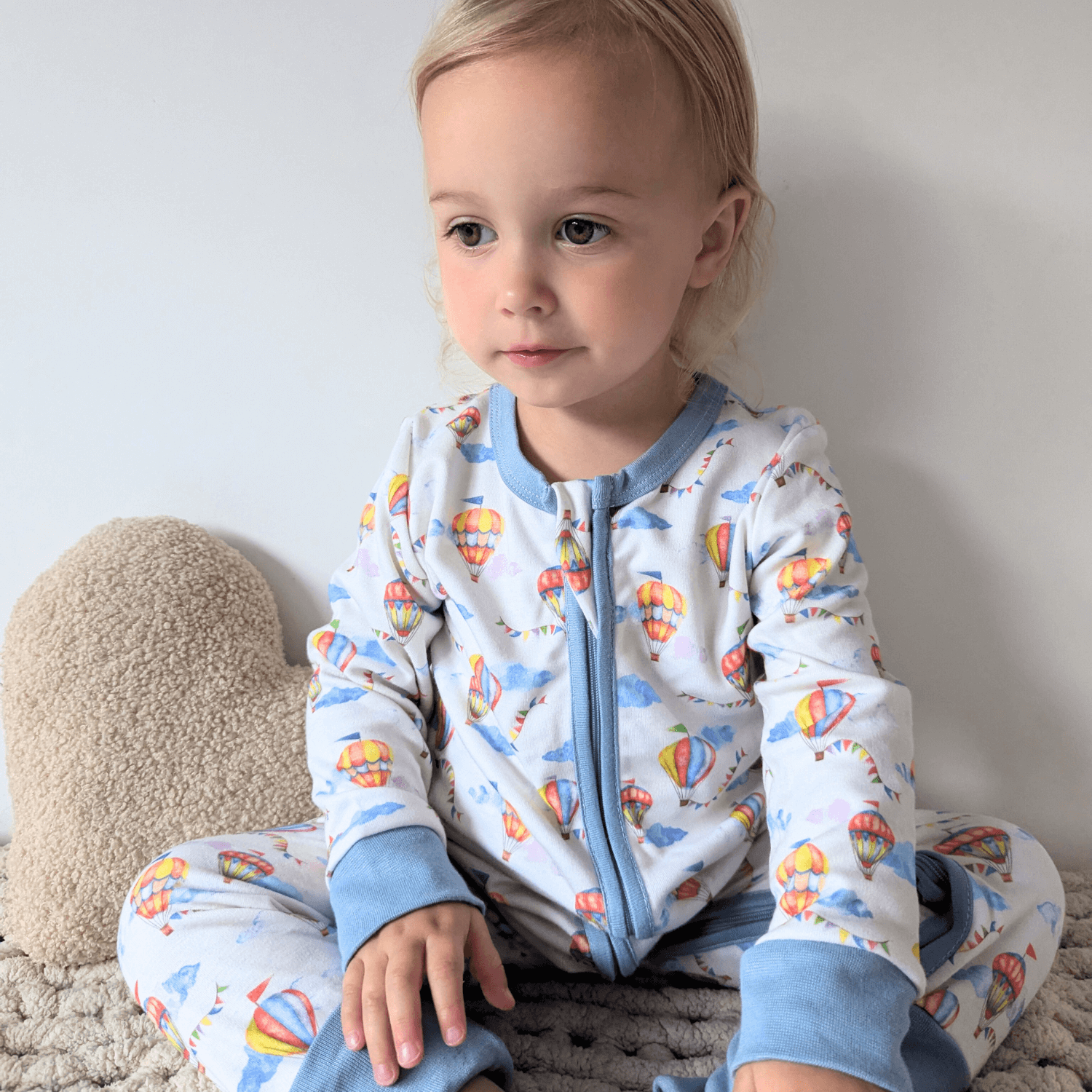 Toddler sat in bed wearing the Up, Up & Away Sleepsuit with hot air balloon and cloud print made from bamboo and organic cotton from GrowGrows