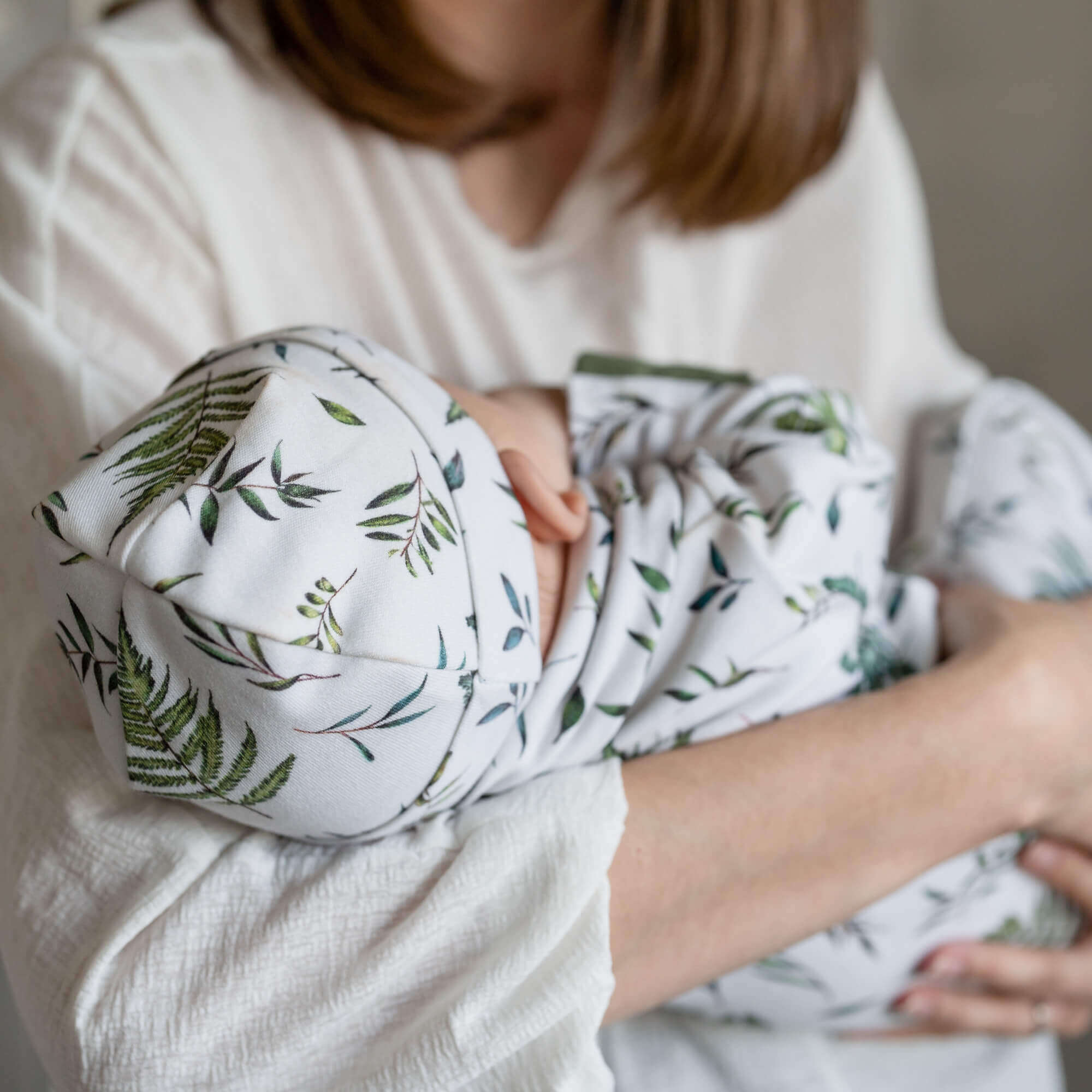 A baby wearing a matching GrowGrows sleepsuit with a matching baby hat and baby bib, held by an adult in a soft cream top. The background is a bright, softly blurred indoor setting with green foliage.