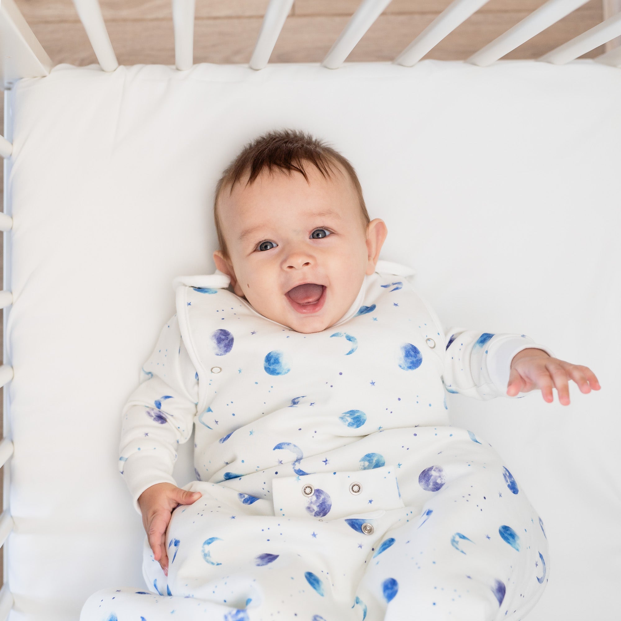 A baby lying in the cot wearing the Marvellous Moons Smart Sleeping Bag 2.5 Tog with blue moon, planet and constellation print made from bamboo and organic cotton from GrowGrows
